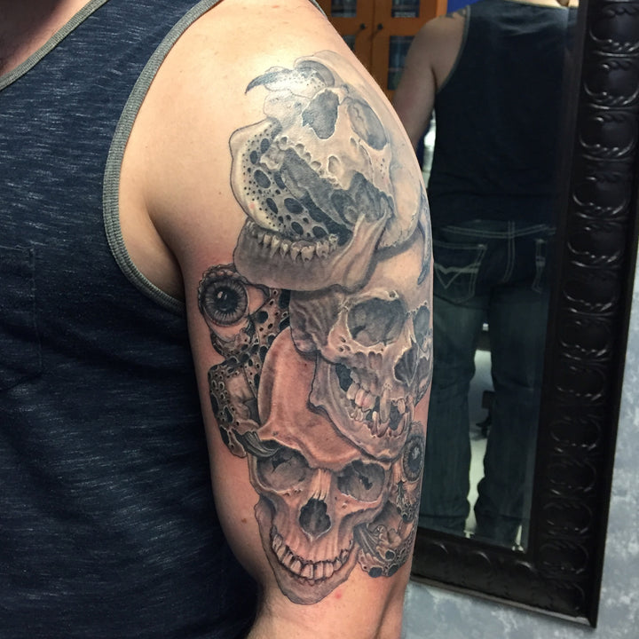 A mans upper arm covered with a tattoo of three realistic black and grey skulls intertwined with an alien creature.