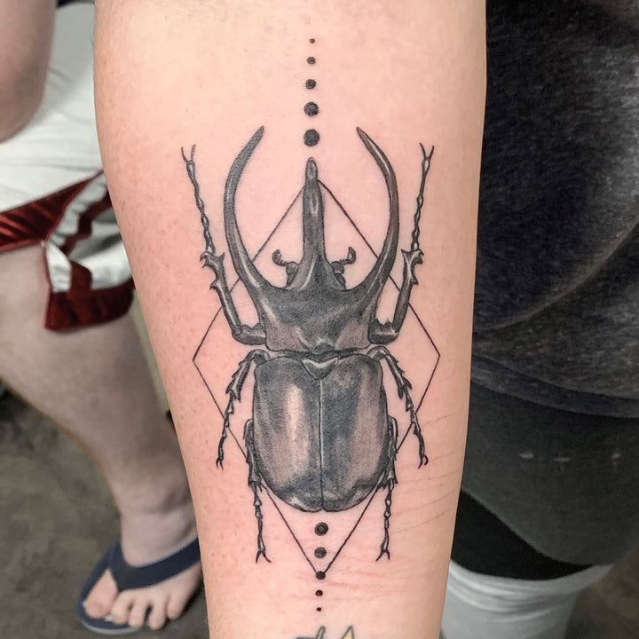 Black and grey Hercules beetle on the inside of a forearm. a geometric diamond outline and dos are in the background