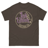 A dark brown t-shirt with a gold circle containing fancy lettering in purple and gold that says Ink Union and a gold tattoo machine peeking out from behind on the right side.  There is a dot work gradient inside the circle, and the words Tattoo Co. in gold are at the bottom of the design.