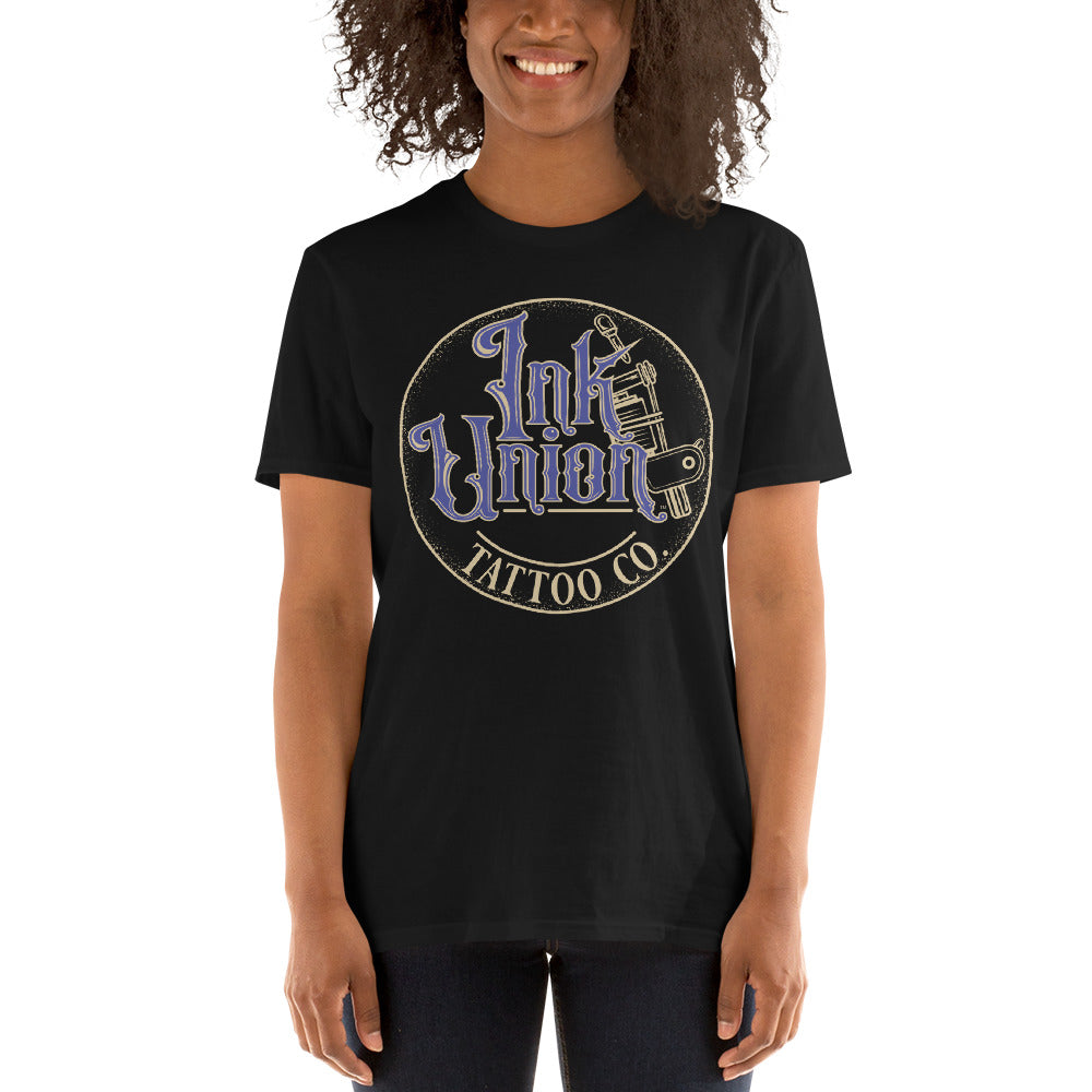 An attractive woman wearing a black t-shirt with a gold circle containing fancy lettering in blue and gold that says Ink Union and a gold tattoo machine peeking out from behind on the right side.  There is a dot work gradient inside the circle, and the words Tattoo Co. in gold are at the bottom of the design.