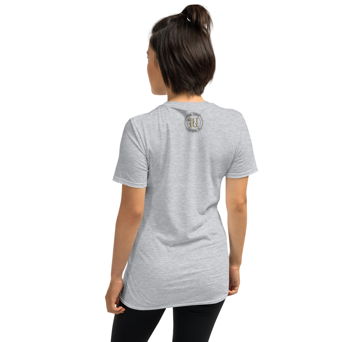 The rear view of an attractive woman wearing a light grey t-shirt with a small gold and black Ink Union badge logo centered just under the neckline. 