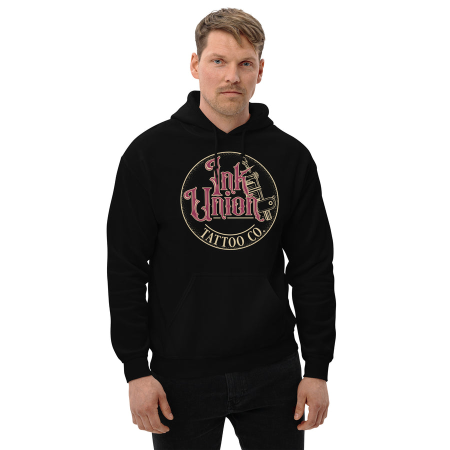 An attractive man wearing a black hoodie with a gold circle containing fancy lettering in red and gold that says Ink Union and a gold tattoo machine peeking out from behind on the right side.  There is a dot work gradient inside the circle, and the words Tattoo Co. in gold are at the bottom of the design.
