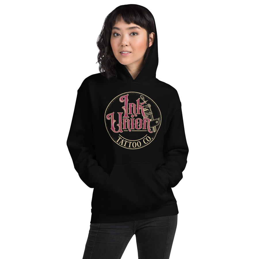 An attractive woman wearing a black hoodie with a gold circle containing fancy lettering in red and gold that says Ink Union and a gold tattoo machine peeking out from behind on the right side.  There is a dot work gradient inside the circle, and the words Tattoo Co. in gold are at the bottom of the design.