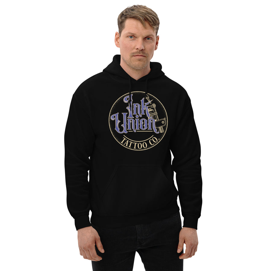 An attractive man wearing a black hoodie with a gold circle containing fancy lettering in blue and gold that says Ink Union and a gold tattoo machine peeking out from behind on the right side.  There is a dot work gradient inside the circle, and the words Tattoo Co. in gold are at the bottom of the design.