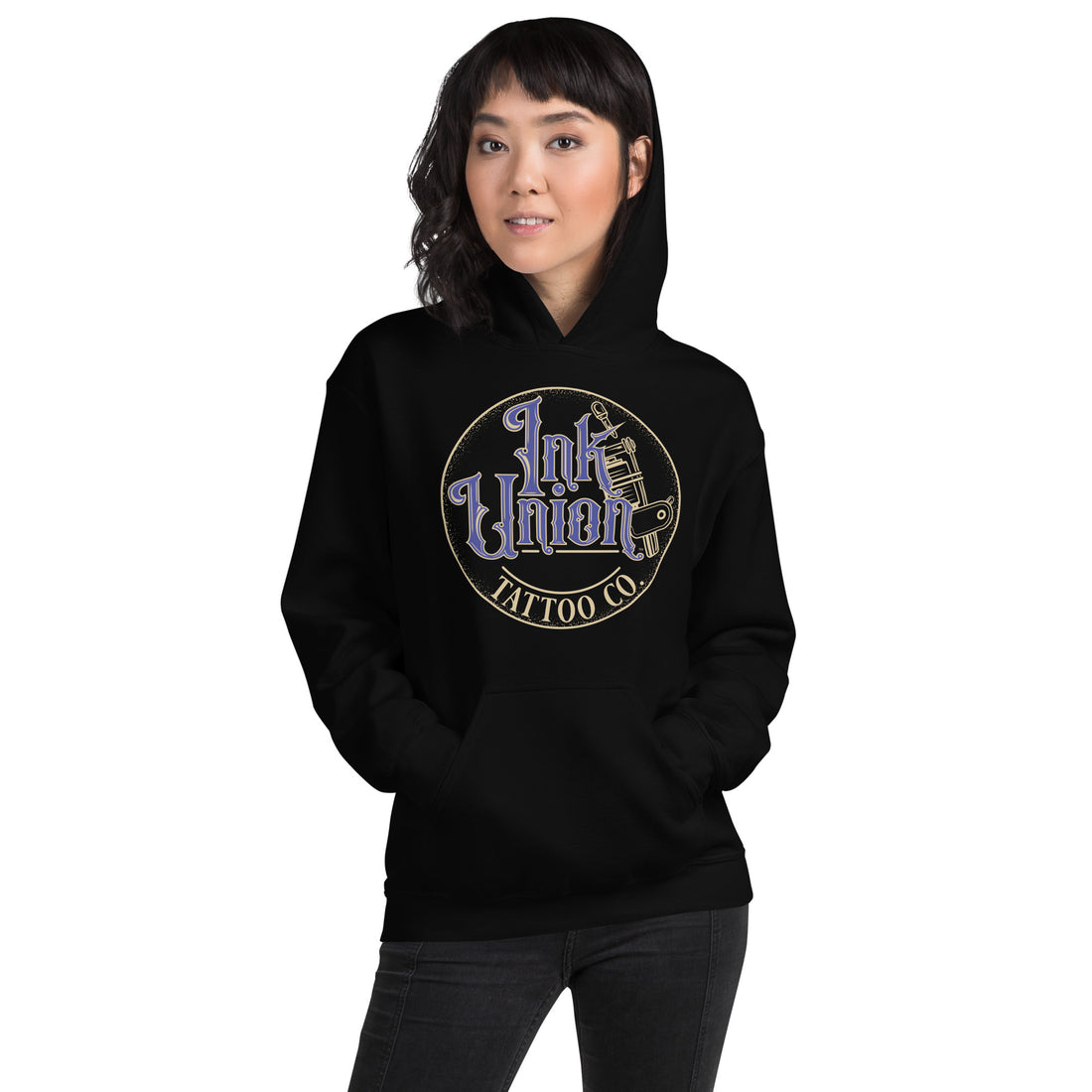 An attractive woman wearing a black hoodie with a gold circle containing fancy lettering in blue and gold that says Ink Union and a gold tattoo machine peeking out from behind on the right side.  There is a dot work gradient inside the circle, and the words Tattoo Co. in gold are at the bottom of the design.